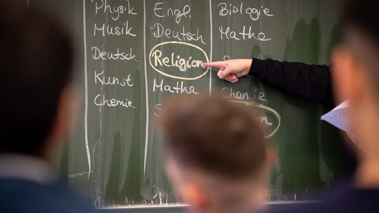 Schülerin Delin steht an einer Tafel, auf der ein Stundenplan mit Religionsunterricht geschrieben ist.