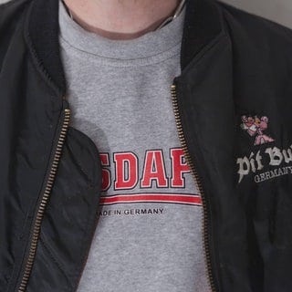 Ein Consdaple-Shirt unter einer geöffneten Bomberjacke. In Neonazikreisen wird diese Kombination gewählt, um die Buchstaben NSDAP aus der Jacke herausblitzen zu lassen.