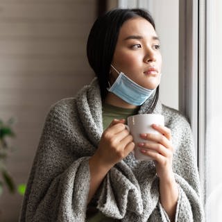 Sick asian girl in medical face mask standing by the window and yearning to go outside, being on quarantine