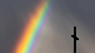 Ein kräftiger Regenbogen ist am dunklen Himmel neben dem Kreuz einer Kirche zu sehen.