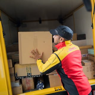 Ein Paketzusteller stellt ein Paket in ein Fahrzeug der Deutschen Post DHL. Es gibt einen neuen Betrugsversuch per Mail. Du wirst aufgefordert eine Zollgebühr für dein Paket zu zahlen.