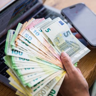 Eine Person hält mehrere tausend Euro in Bargeld in der Hand, dahinter steht ein Laptop und sie hält ihr Handy in der Hand.