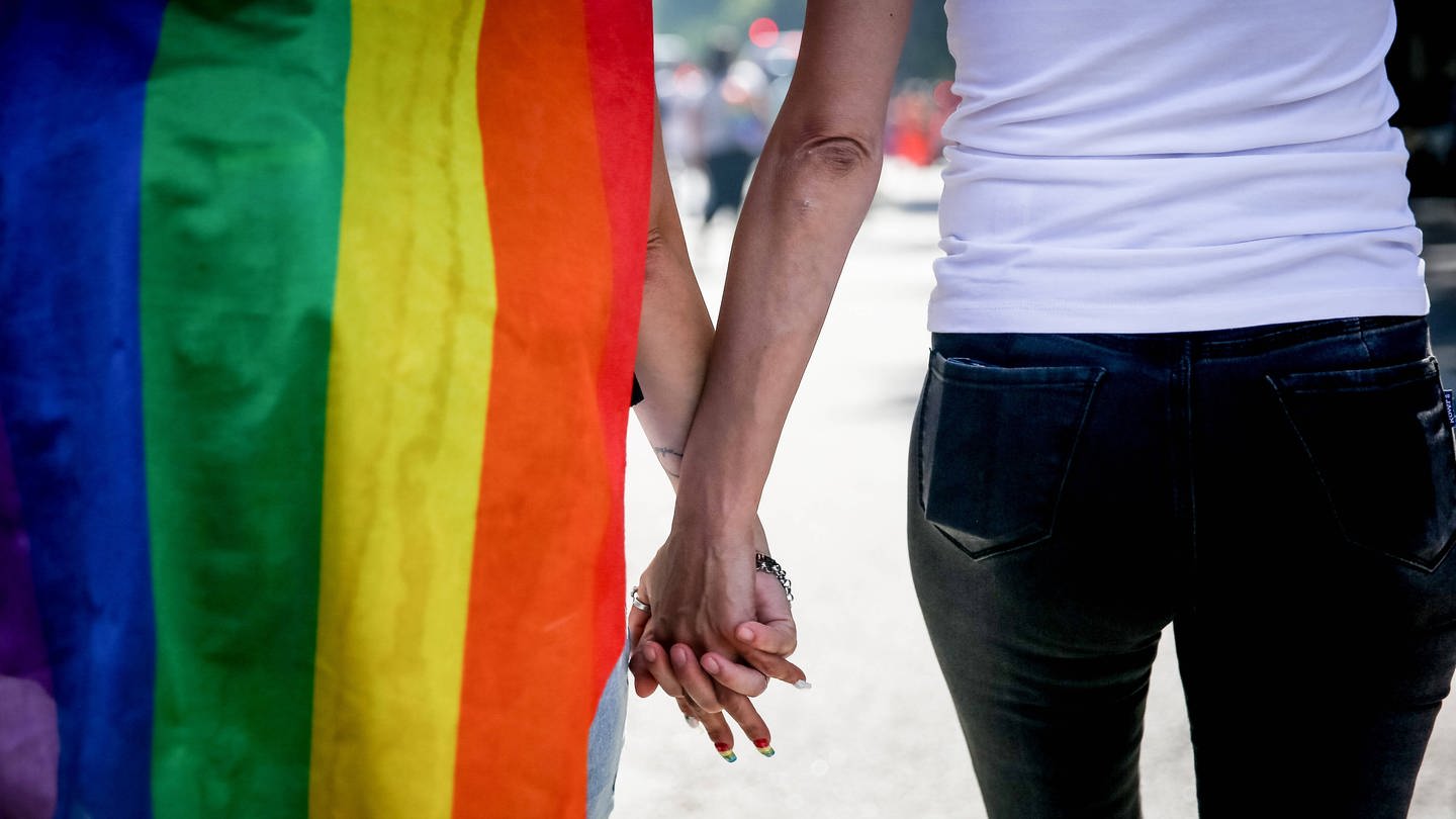 Zwei Personen halten Händchen und haben eine Regenbogen-Flagge umhängen.