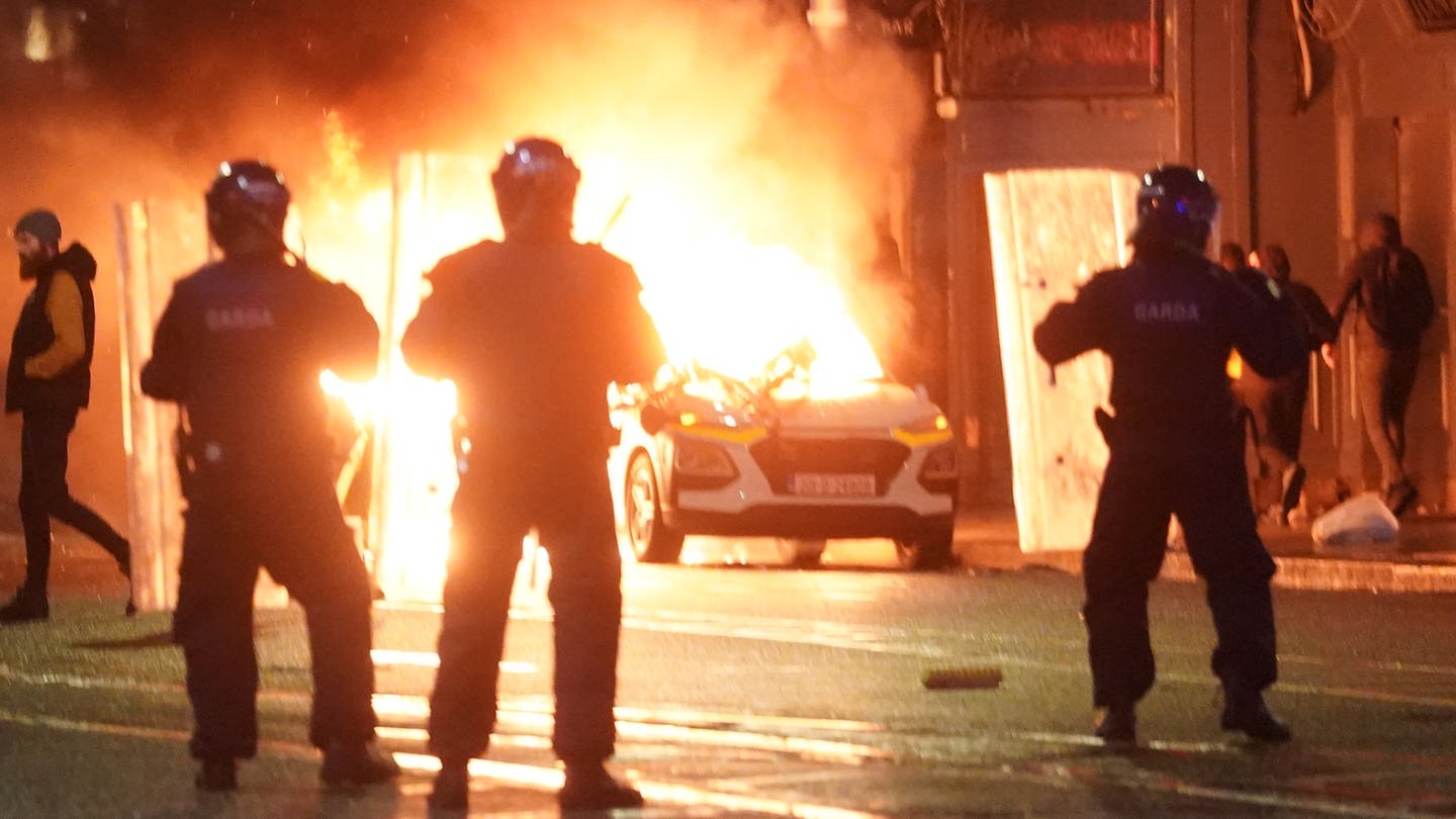 Irische Nationalpolizisten stehen in der Nähe eines brennenden Polizeiautos. Nach einem Messerangriff in der irischen Hauptstadt, bei dem unter anderem ein fünfjähriges Mädchen und eine Frau schwer verletzt wurden, kam es am Abend in der Nähe des Tatorts zu schweren Ausschreitungen.