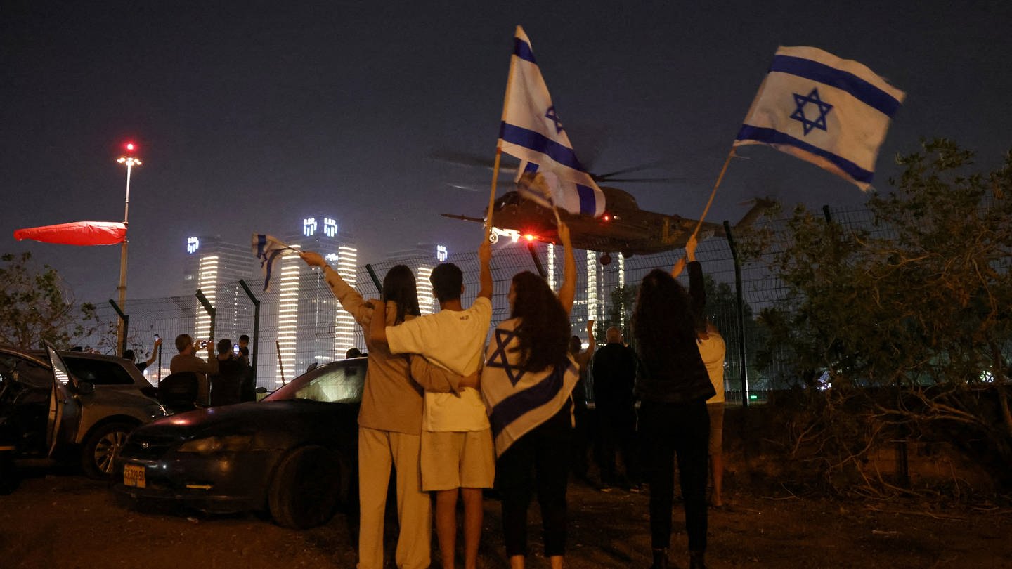 Israelis erwarten mit schwenkenden Fahnen die Ankunft der Geiseln in einem Helikopter.