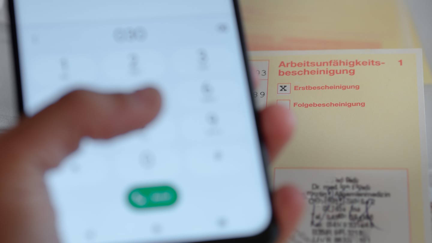 Eine Person hält ein Smartphone in der Hand, daneben liegt eine Krankschreibung.