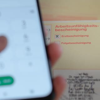 Eine Person hält ein Smartphone in der Hand, daneben liegt eine Krankschreibung.