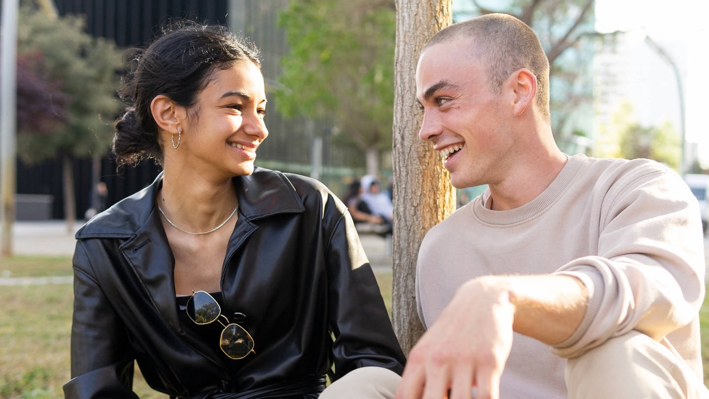 SYMBOLBILD: Eine junge Frau und ein junger Mann sitzen gemeinsam in der Wiese und lachen einander an.
