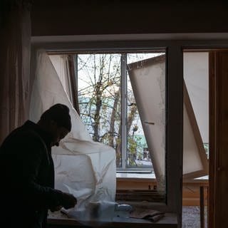 Ein Mann deckt ein zerbrochenes Fenster in seiner Wohnung nach einem russischen Drohnenangriff mit Plastik ab. 