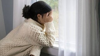 Depression: Eine junge Frau steht am Fenster und hält ihre Händen über das Gesicht (Symbolbild)