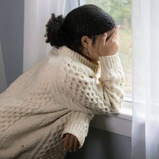 Depression: Eine junge Frau steht am Fenster und hält ihre Händen über das Gesicht (Symbolbild)