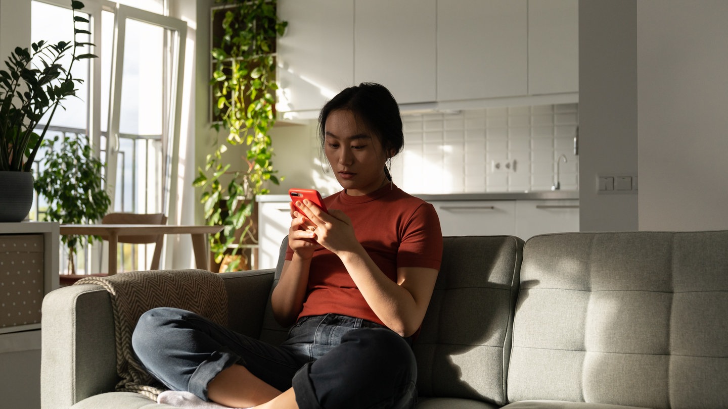 Eine junge Frau sitzt auf dem Sofa und guckt auf ihr Handy.