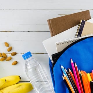 Symbolbild: Eine Schultasche mit Wasser, Banane, Nüssen und Stiften.