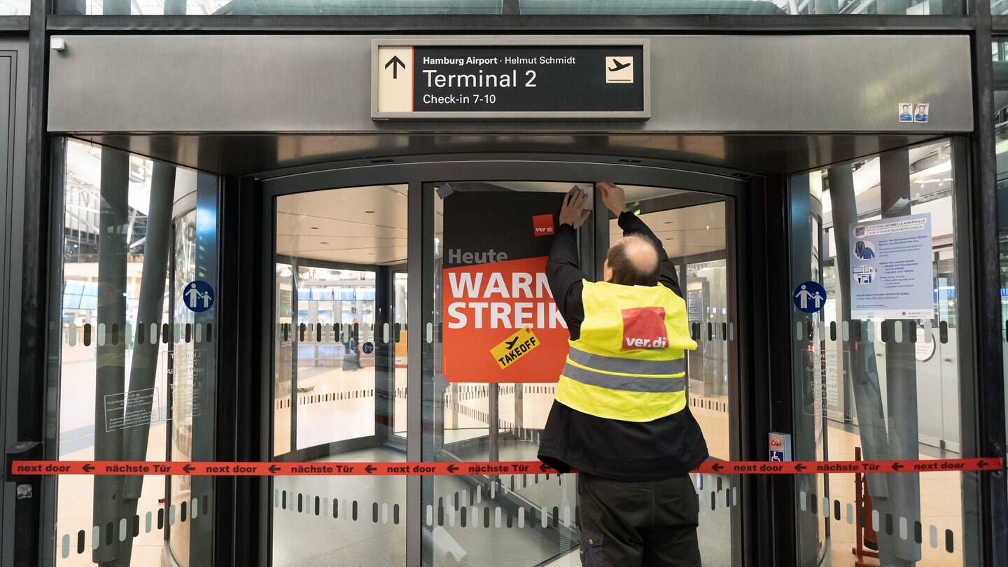 Ein Verdi-Vertreter hängt ein Plakat mit der Aufschrift «Warnstreik» an einer geschlossenen Terminaldrehtür am Hamburger Flughafen auf.