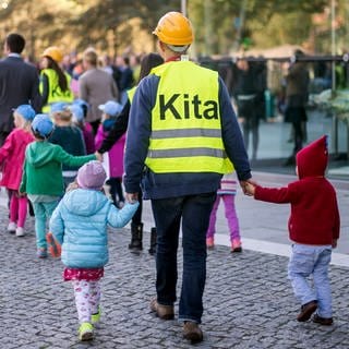 Kinder der Kindertagesstätte "Wolkenzwerge" laufen am 01.10.2015 in Berlin an den Händen eines Betreuers. Dieser trägt eine gelbe Warnweste, auf die der Schriftzug "Kita" gedruckt ist.