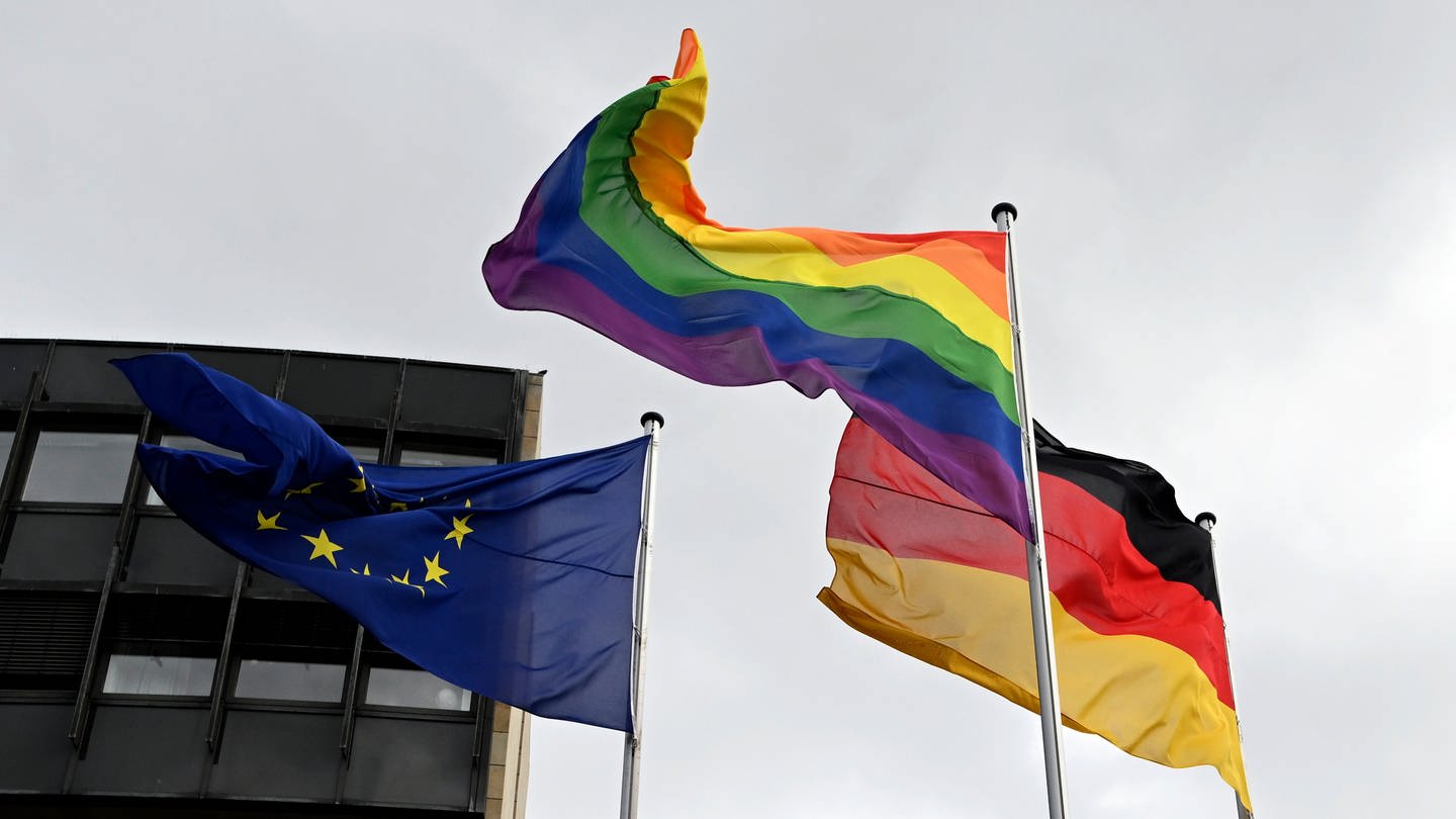Eine Regenbogenflagge weht neben einer Deutschlandflagge und einer Europaflagge