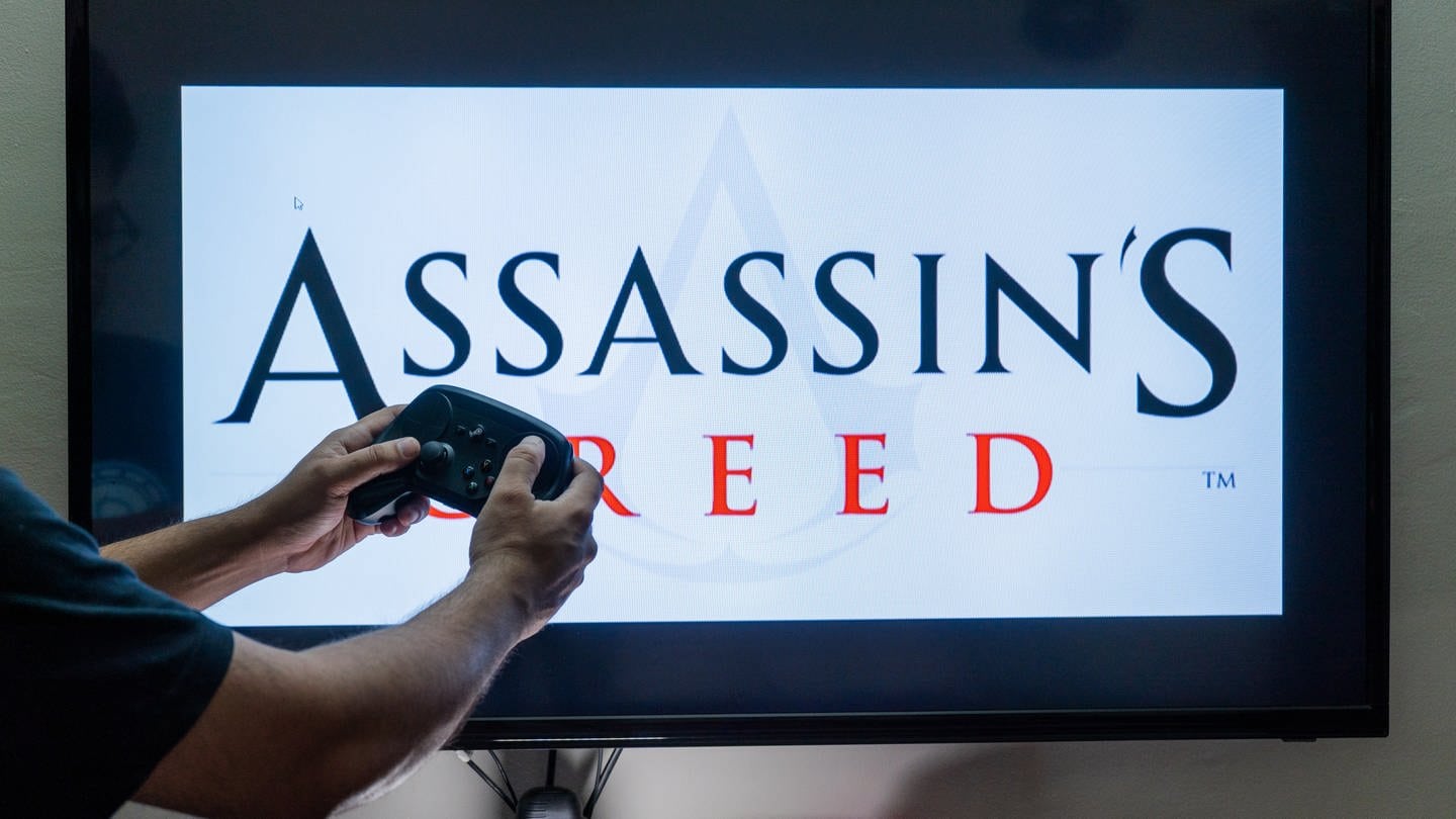 Man holding steam controller in front of a screen loading a game of assassin s creed an action adventure game by ubisoft with multiple installements
