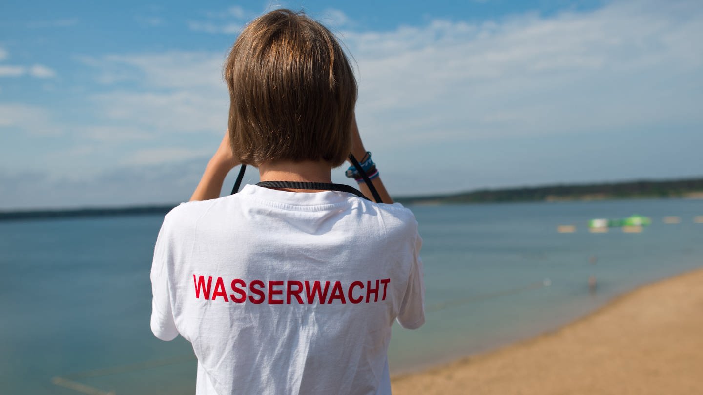 Eine Rettungsschwimmerin von der Wasserwacht schaut durch ihr Fernglas auf einen See - Im Pfuhler See in Neu-Ulm ist fast ein 28-Jähriger ertrunken