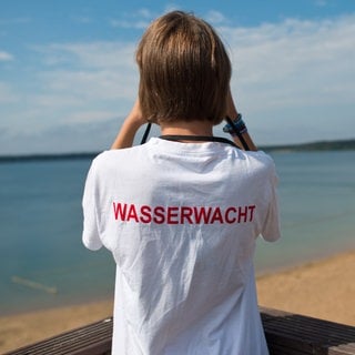 Eine Rettungsschwimmerin von der Wasserwacht schaut durch ihr Fernglas auf einen See - Im Pfuhler See in Neu-Ulm ist fast ein 28-Jähriger ertrunken