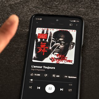 Finger vor Smartphone mit Musik App streamt das Lied "L'amour toujours" von Gigi D'Agostino. 