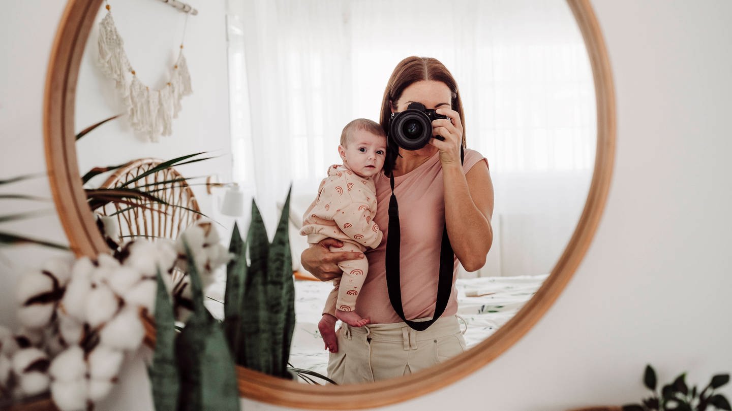 Mutter mit Baby auf dem Arm macht Selfie vor einem Spiegel.