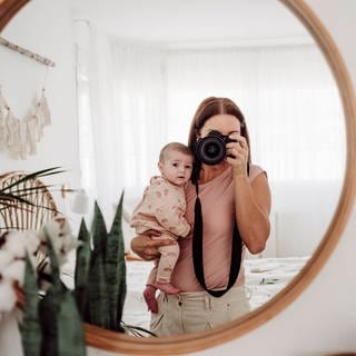 Mutter mit Baby auf dem Arm macht Selfie vor einem Spiegel.