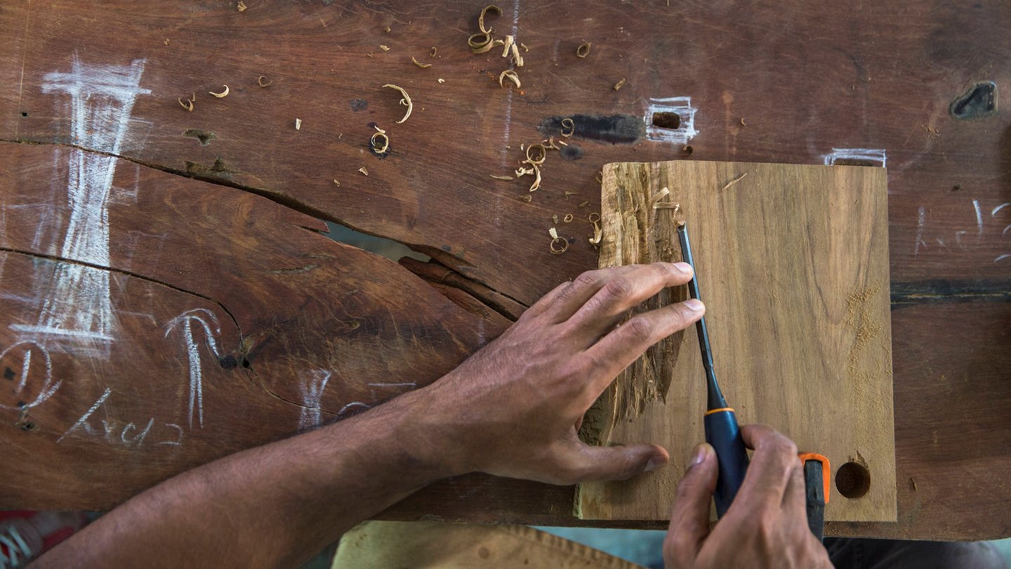 Die Hände eines Zimmermanns schnitzen eine Holzplatte auf einem Arbeitstisch.