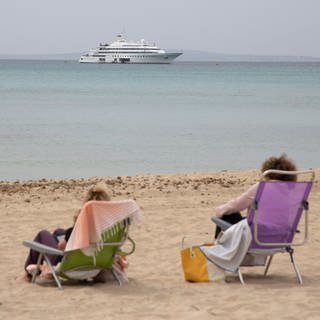 SYMBOLBILD - Frauen sitzen an einem Strand. Auf dem Meer ist eine Jacht zu sehen.