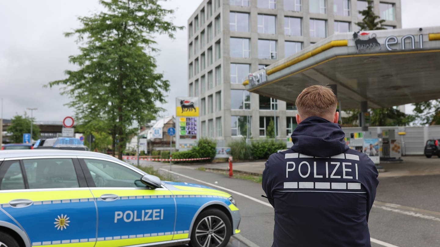 Ein Bereich nahe einer Tankstelle ist mit Absperrband gesichert, davor ist ein Polizeiwagen und ein Polizist zu sehen. Nach mehreren Schüssen auf eine Menschengruppe an einer Tankstelle in Konstanz ist ein Mann mit schweren Verletzungen in ein Krankenhaus gekommen.