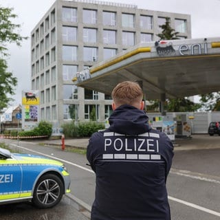 Ein Bereich nahe einer Tankstelle ist mit Absperrband gesichert, davor ist ein Polizeiwagen und ein Polizist zu sehen. Nach mehreren Schüssen auf eine Menschengruppe an einer Tankstelle in Konstanz ist ein Mann mit schweren Verletzungen in ein Krankenhaus gekommen. 