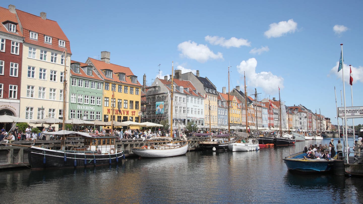 Passanten schlendern im Nyhavn am Wasser entlang. Der Nyhavn mit seinen bunten Häuschen zählt zu den größten Sehenswürdigkeiten von Kopenhagen.