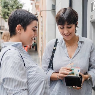 Zwei junge Frauen stehen vor einem Bankautomat und heben Bargeld ab.