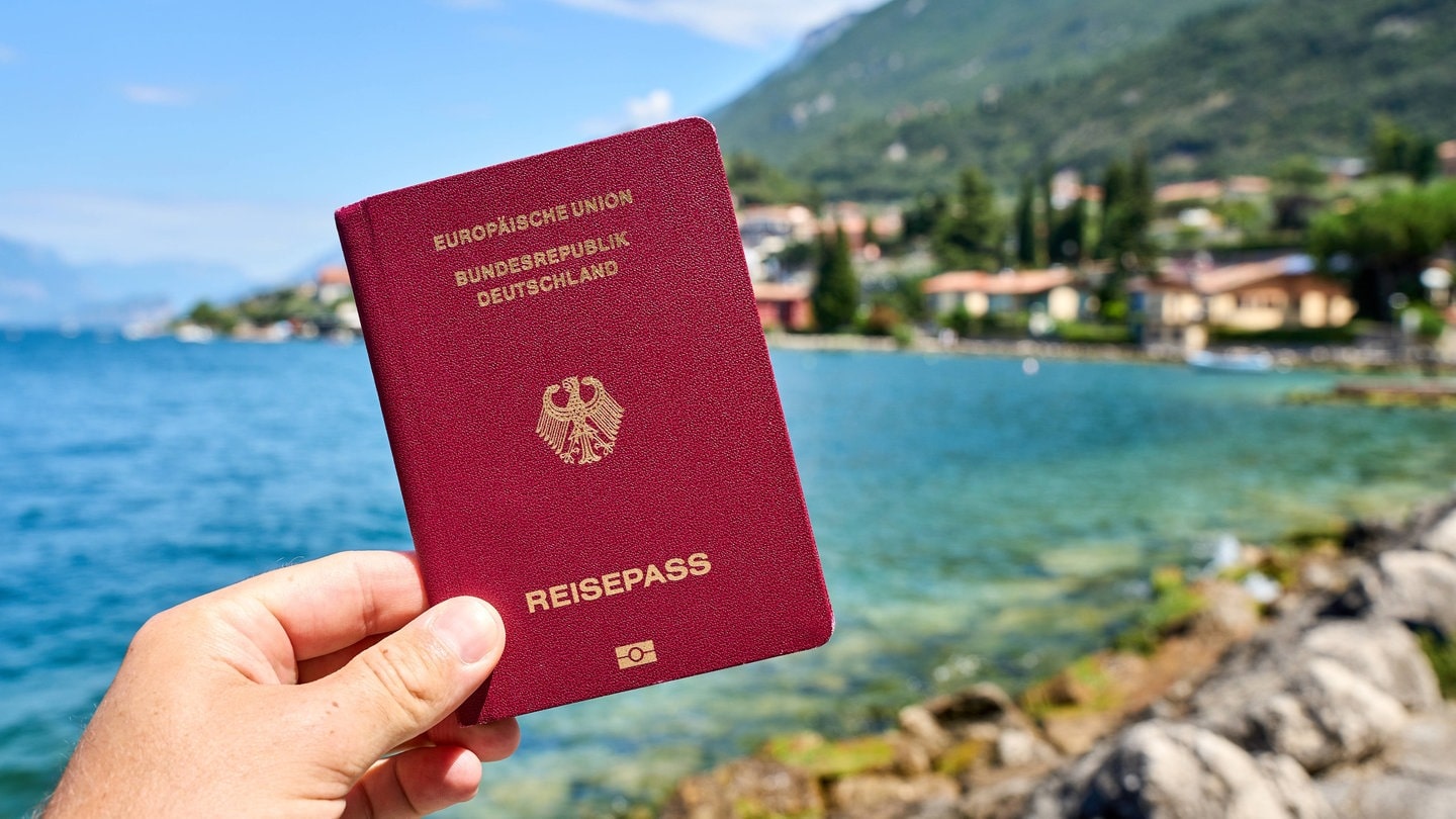 Ein Mann hält den deutschen Reisepass. Im Hintergrund ist der Gardasee in Italien zu sehen.