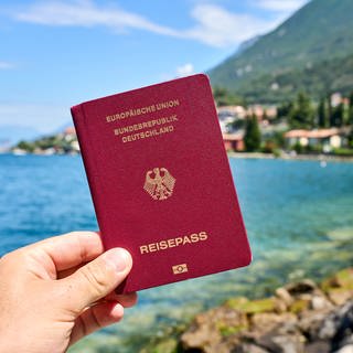 Ein Mann hält den deutschen Reisepass. Im Hintergrund ist der Gardasee in Italien zu sehen.