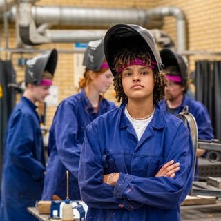 Junger Mann mit Locken im Blaumann. Eine Gruppe junger Menschen im Blaumann hinter ihm arbeiten an einer Maschine. 