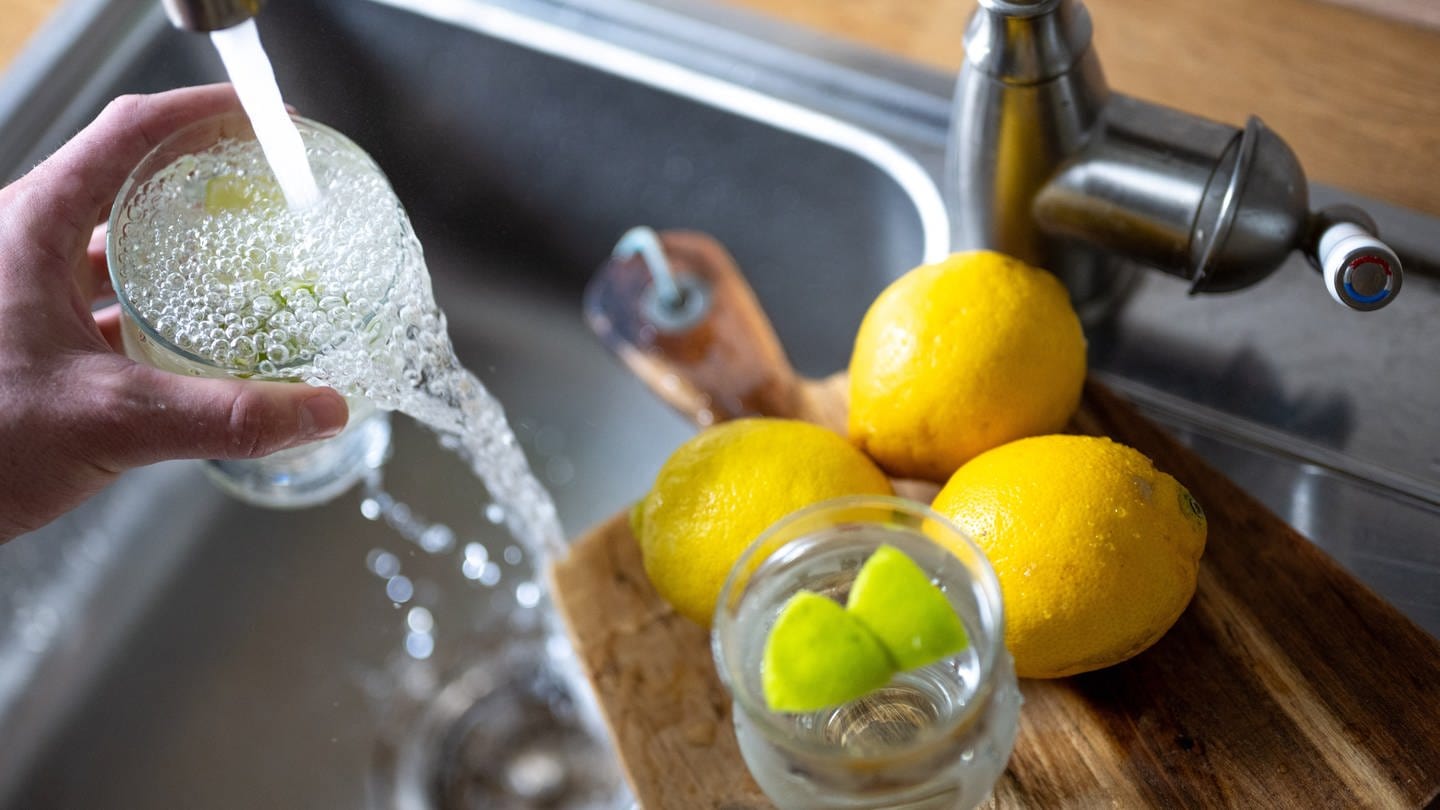 Tinkwasser läuft aus dem Wasserhahn in einer Küche in ein Glas mit Limettenstücken, während im Vordergrund Zitronen auf einem Küchenbrett liegen.