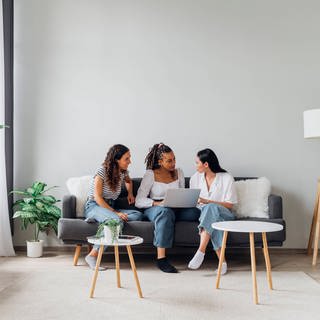 SYMBOLBILD: Drei Frauen sitzen auf einer Couch in einem WG-Zimmer und unterhalten sich.