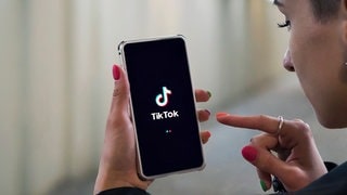 Eine junge Frau hält ihr Handy in den Händen mit einem TikTok-Logo auf dem Bildschirm (Symbolbild)