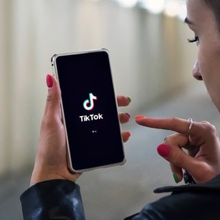 Eine junge Frau hält ihr Handy in den Händen mit einem TikTok-Logo auf dem Bildschirm (Symbolbild)