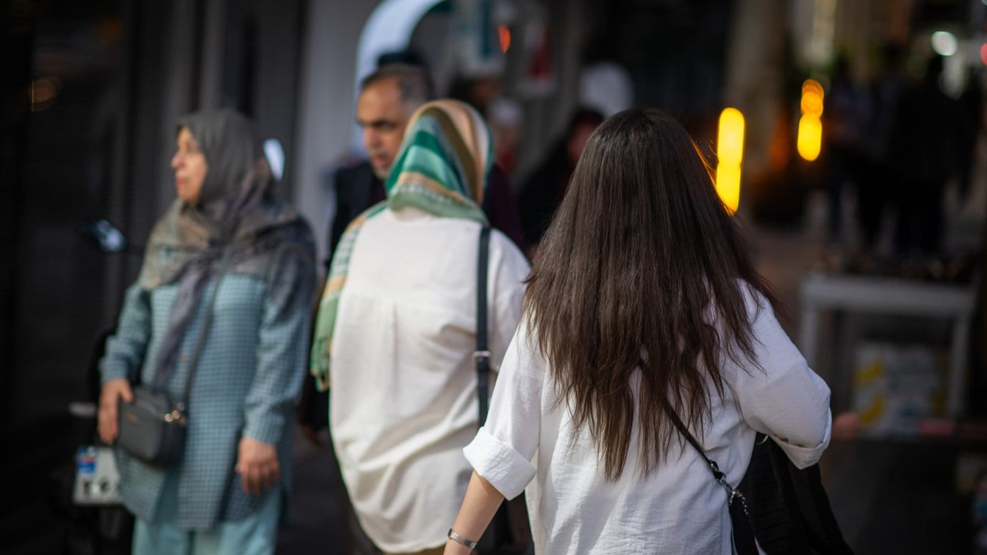 Eine Frau läuft in Teheran mit offenen Haaren am Abend eine Straße entlang, im Hintergrund laufen zwei Frauen mit Kopftuch.