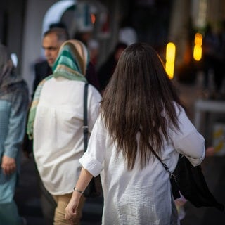 Eine Frau läuft in Teheran mit offenen Haaren am Abend eine Straße entlang, im Hintergrund laufen zwei Frauen mit Kopftuch.