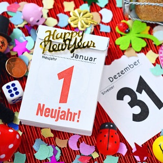 Kalenderblätter vom 31. Dezember und 1. Januar mit Aufschrift "Neujahr!" umgeben von Glückssymbolen und Konfetti.