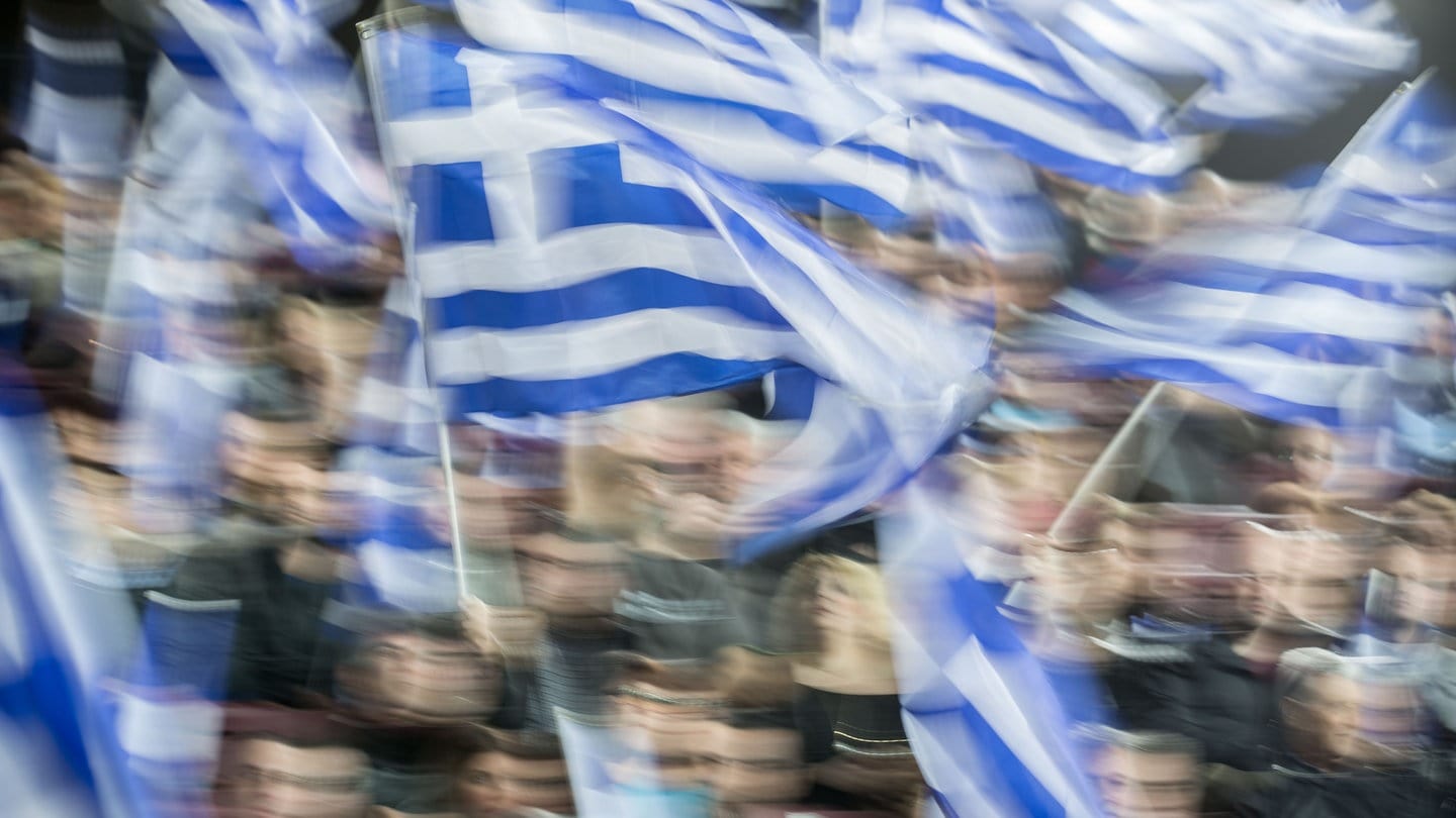 Menschen mit Griechenland-Fahnen (Symbolbild) - Zum Jahrestag des Zugunglücks in Griechenland wird mit Ausschreitungen