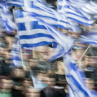 Menschen mit Griechenland-Fahnen (Symbolbild) - Zum Jahrestag des Zugunglücks in Griechenland wird mit Ausschreitungen